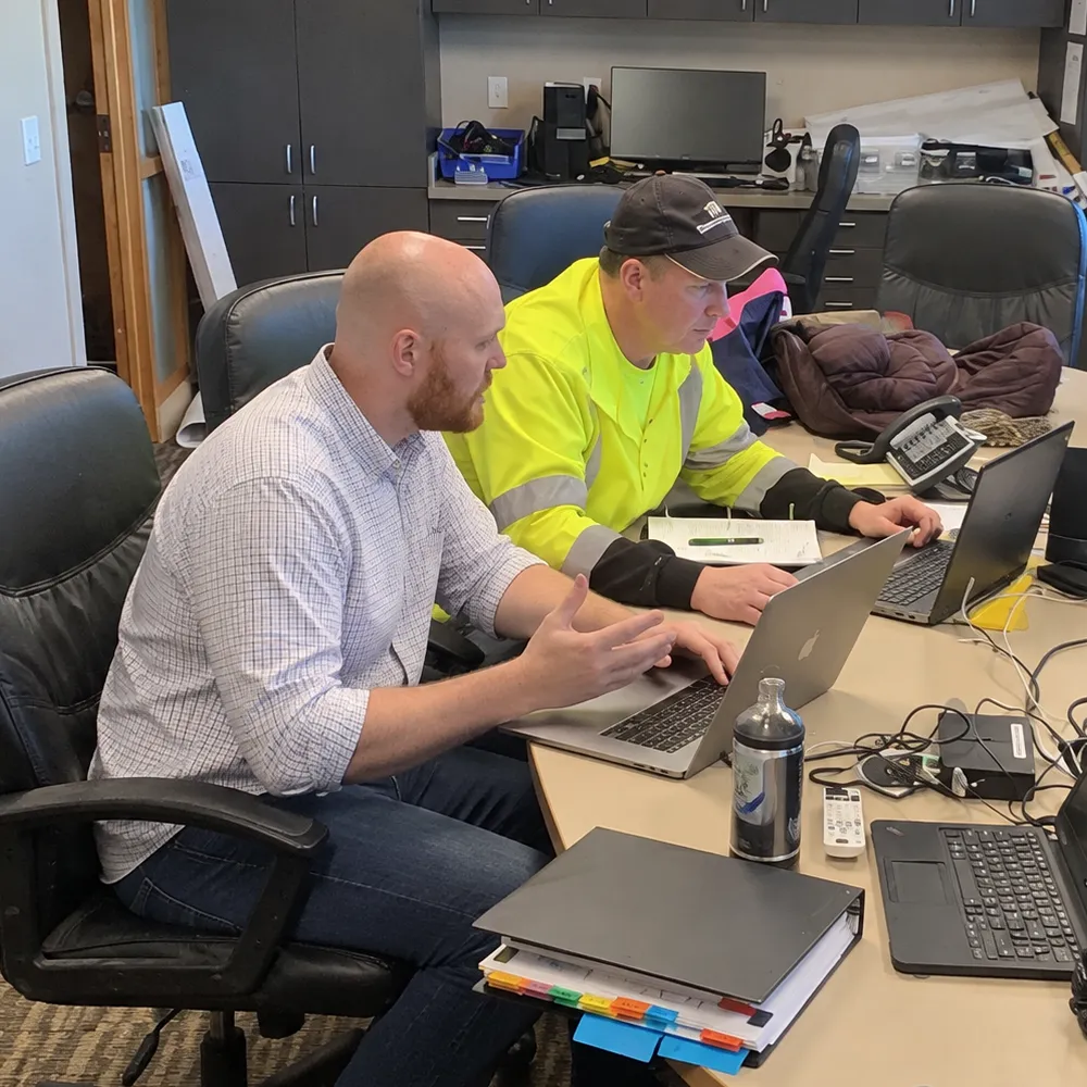 Two estimators at a conference table, one talking through a bid on his laptop while the other, in a hi-vis shirt, reads along.