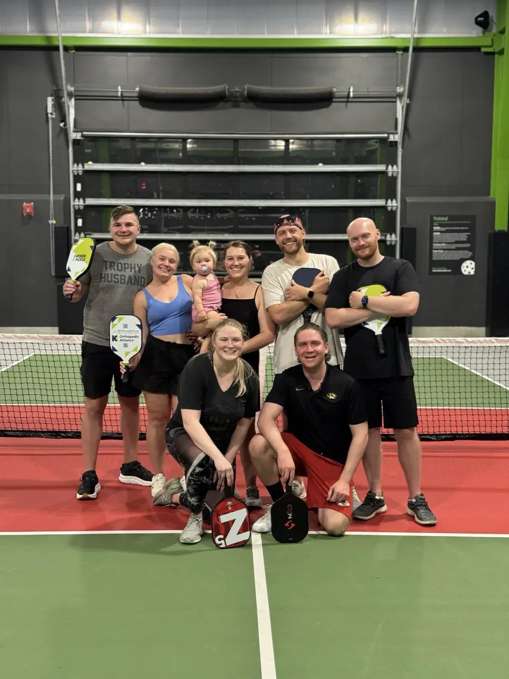 Ocho del equipo y un niño pequeño en una cancha techada de pickleball, raquetas en mano.