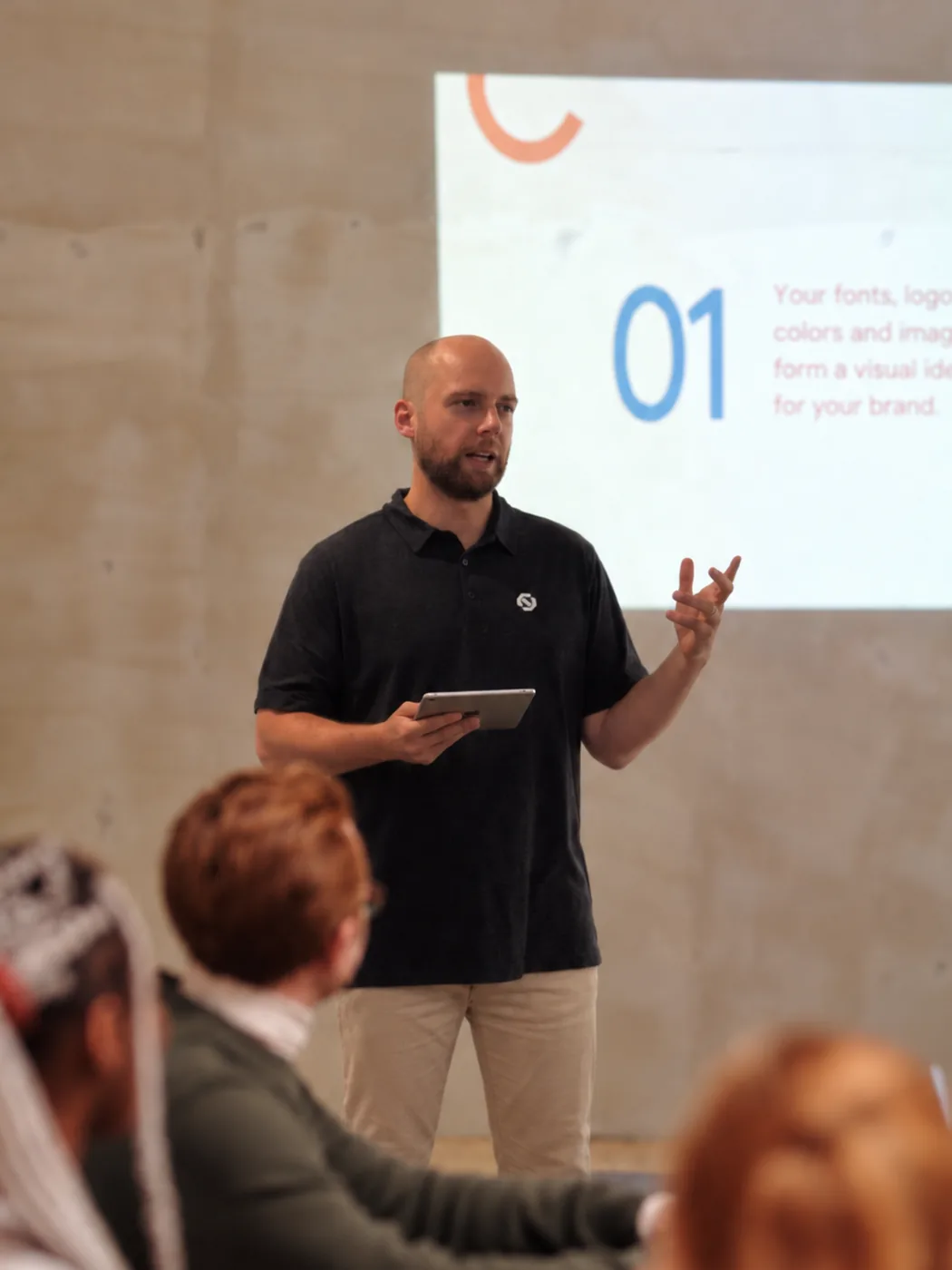 A teammate presenting to the room, tablet in hand, with a slide behind them.